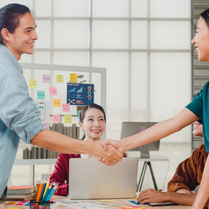 Multiracial group of young creative people in smart casual wear discussing business shaking hands together and smiling while standing in modern office. Partner cooperation, coworker teamwork concept.